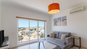 A modern living room with a sofa, a table, a chair, a TV, and a large window with a view of buildings. - Quinta dos Arcos Lote 18 3ºG Apartment with Sea View, Pool and Balcony (Armação de Pêra)