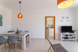 Un salon moderne comprenant un coin repas, un téléviseur et une cuisine visible à travers une porte ouverte.