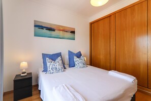 Un lit bien fait, avec du linge de lit blanc, une table de chevet avec une lampe et, au mur, un tableau représentant un coucher de soleil sur l’eau.