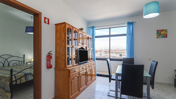 A room with a bed, a wooden cabinet with a television, a dining table with chairs, a window with curtains, and a fire extinguisher on the wall. - Torre Sol 909 Beach Apartment with Pool, Garden, and Balcony (Armação de Pêra)