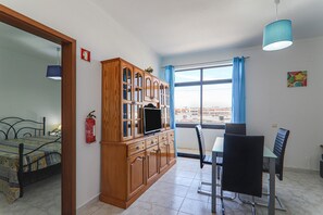 Um quarto com uma cama, um armário de madeira com uma televisão, uma mesa de jantar com cadeiras, uma janela com cortinas e um extintor de incêndio na parede.