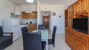 A compact kitchen with a refrigerator, a dining table with chairs, a television on a wooden cabinet, and a blue hanging light fixture. - Torre Sol 909 Beach Apartment with Pool, Garden, and Balcony (Armação de Pêra)