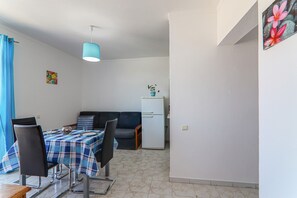 Uma sala de jantar com mesa posta para dois, um sofá, um refrigerador e uma luminária pendente azul.