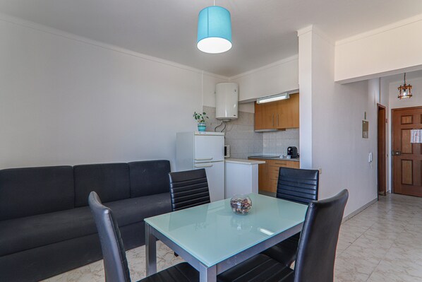 Espaço compacto com sofá, mesa de jantar, cadeiras, pequena cozinha e uma porta que dá para outro cômodo.