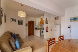 A living room with a sofa, a wooden dining table, and chairs. There is a kitchen area with appliances and a fire extinguisher.