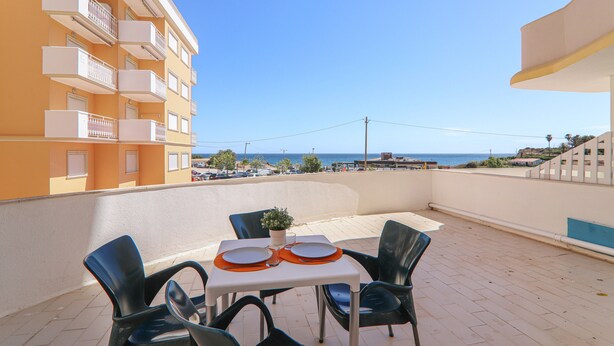 Een balkon met een tafel gedekt voor twee personen, stoelen, een bloempot en uitzicht op de zee en gebouwen.