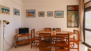 Een eethoek met een ronde houten tafel, zes stoelen, een televisie op een meubel en kunstwerken aan de muur.