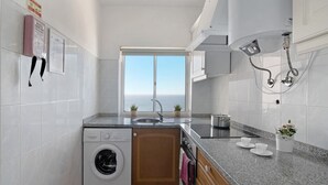 A compact kitchen with a washing machine, a sink, and a window offering a view of the sea.