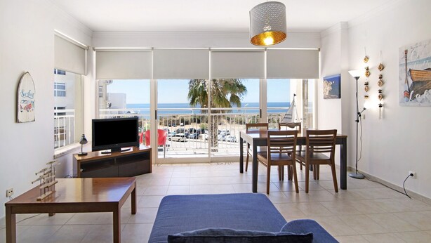 A living room with a TV, a sofa, a dining table with chairs, and a balcony with a view of the sea.