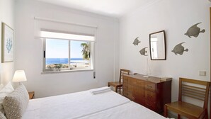 A bedroom with a bed, a desk, a chair, a dresser, a window with a view, and wall art.