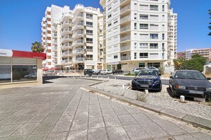 A multi-story residential building with balconies, a parking area with cars, and a building with a red awning.