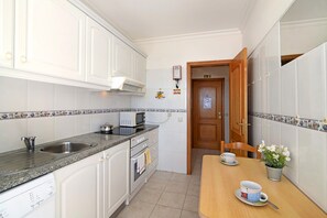 Une cuisine compacte avec des meubles blancs, un four à micro-ondes, un évier et une table à manger en bois, sur laquelle se trouvent une tasse et une soucoupe.