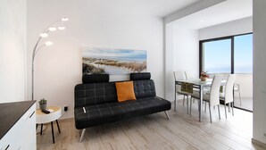 A modern living room with a sofa, dining table, and chairs, a painting on the wall, and a view through a large window. - Apt. Torre Sol 10th 6 - Sea View, A/C and Pool (Armação de Pêra)