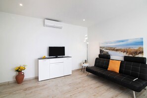 Un salon moderne avec un meuble blanc, un canapé noir agrémenté d’un coussin orange, un téléviseur à écran plat, une plante en pot et un grand tableau représentant un paysage accroché au mur.