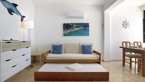 Un salon moderne comprenant un canapé, une table basse, une table à manger avec des chaises, un climatiseur fixé au mur et un tableau représentant un paysage côtier.