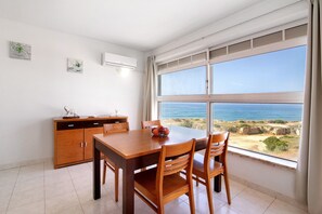 Un coin repas avec une table et des chaises en bois, un petit meuble et une vue sur la mer grâce à de grandes fenêtres.
