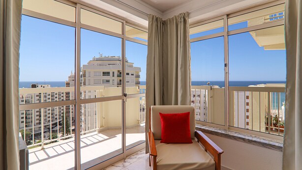 A balcony with a chair and a red pillow, large windows, and a view of the sea and buildings. - Oura 11th Floor Beach Apartment with Sea View, Pool, A/C, Wi-Fi, Garden & Balcony (Armação de Pêra)
