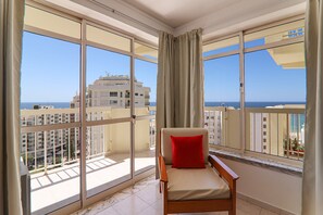 A balcony with a chair and a red pillow, large windows, and a view of the sea and buildings. - Oura 11th Floor Beach Apartment with Sea View, Pool, A/C, Wi-Fi, Garden & Balcony (Armação de Pêra)
