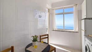 Una cocina pequeña con lavadora, una mesa con planta en maceta y dos vasos de zumo de naranja, y una ventana con vista al mar.