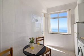 A small kitchen with a washing machine, a table with a potted plant and two glasses of orange juice, and a window with a view of the sea.