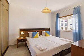 A hotel room with a double bed, a nightstand with a lamp, a wooden headboard, a window with blue curtains, and a view of buildings outside.