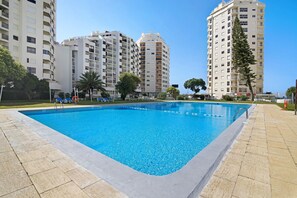 A swimming pool surrounded by high-rise buildings, a clear sky, and some greenery.