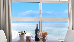 Una mesa con una botella de vino, dos copas, un florero con flores y un bol de naranjas junto a una ventana con vistas al mar.