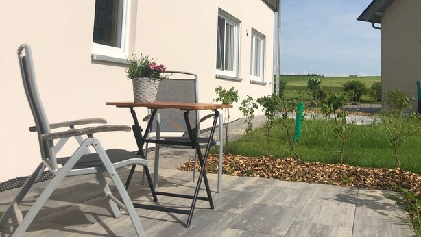 Ein modernes Haus mit Terrasse, einem Tisch, einem Stuhl und einem Blumentopf.