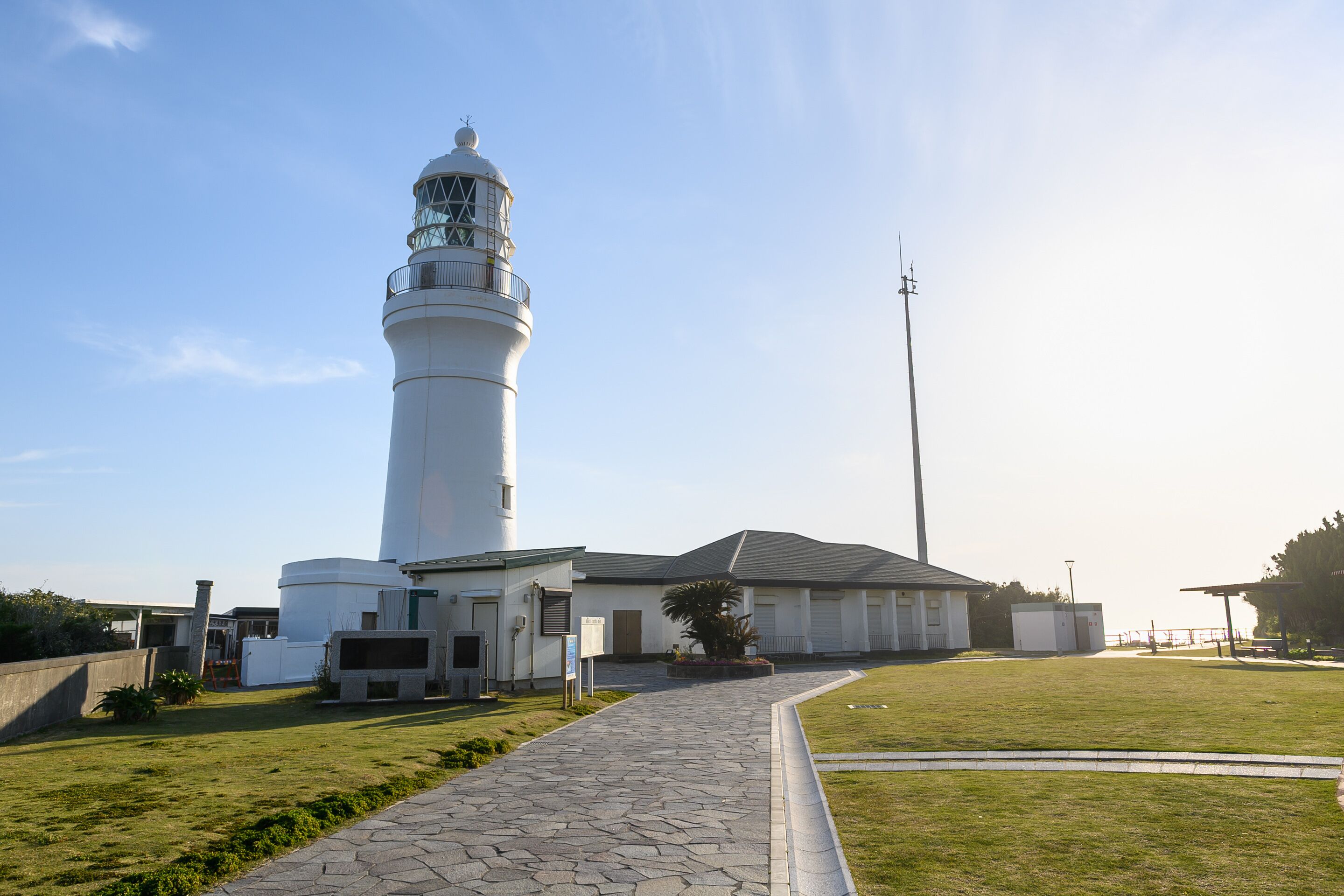 A lighthouse with a pathway leading to its entrance, surrounded by a grassy area and a building with a sloped roof.