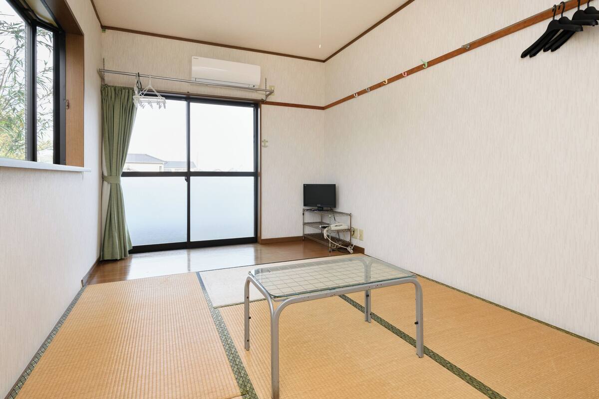 Ein Zimmer mit Tatami-Boden, einem Futon-Bett, einem Fernseher auf einem Standfuß und einer Schiebetür mit Blick hinaus.