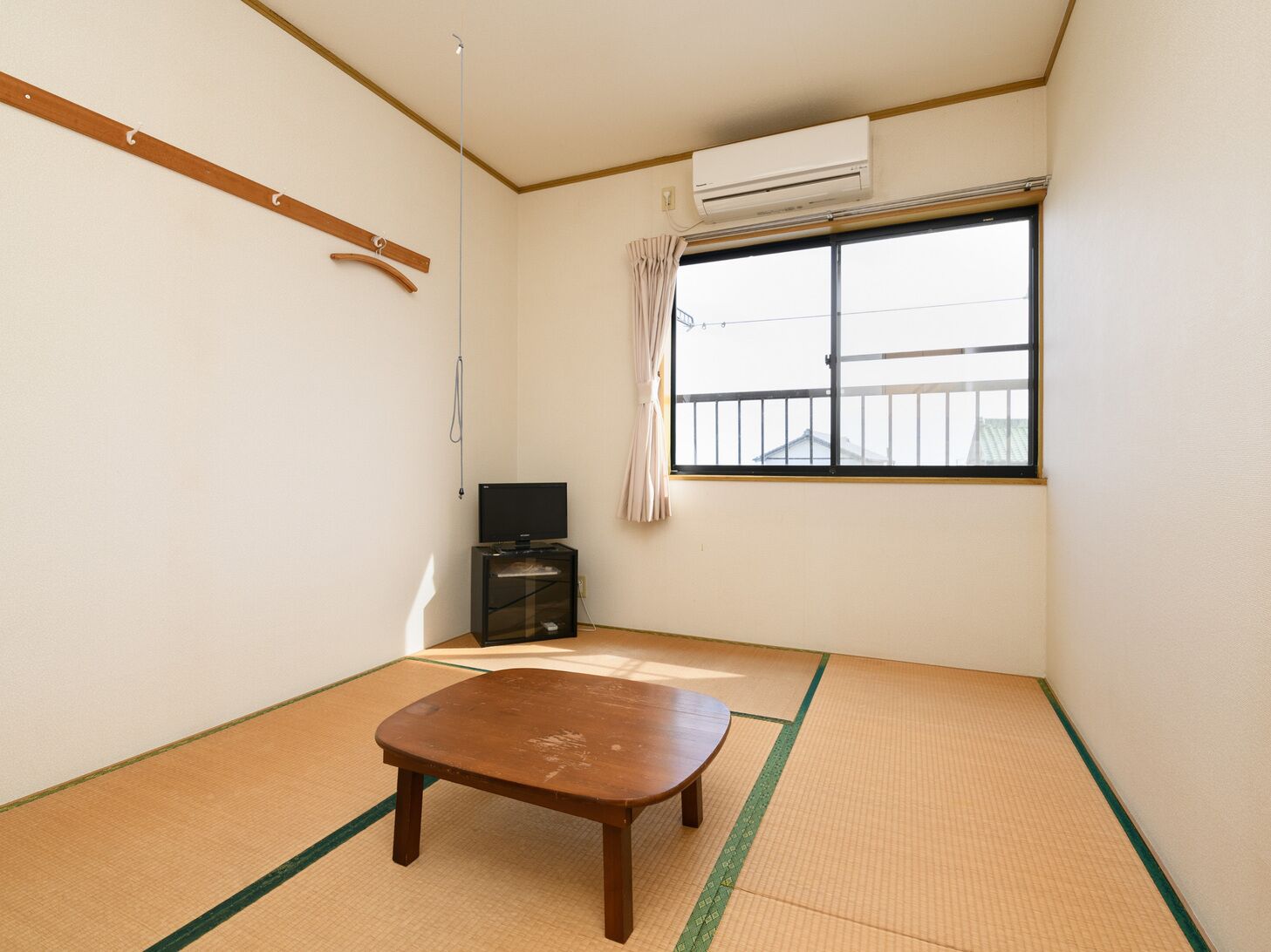 Ein Zimmer mit Tatami-Böden, einem Fernseher, einem kleinen Schrank und einem Fenster mit Blick auf ein Gebäude.