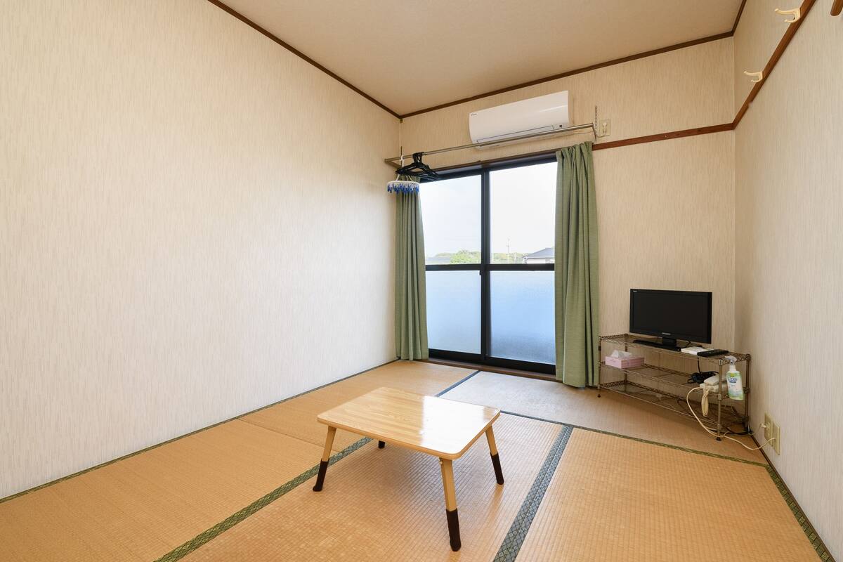 Ein Zimmer mit Tatami-Boden, einem Holztisch, einem Fernseher auf einem Standfuß und Meerblick durch eine Schiebetür.