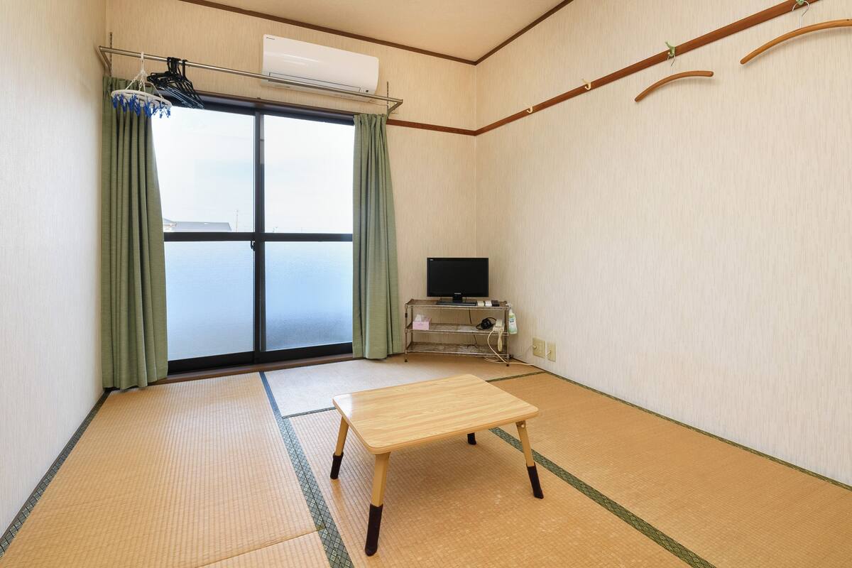 Ein Zimmer mit Tatami-Boden, einem Fernseher auf einem Schrank, einem Fenster mit Vorhängen und einem Holzbalken an der Wand.