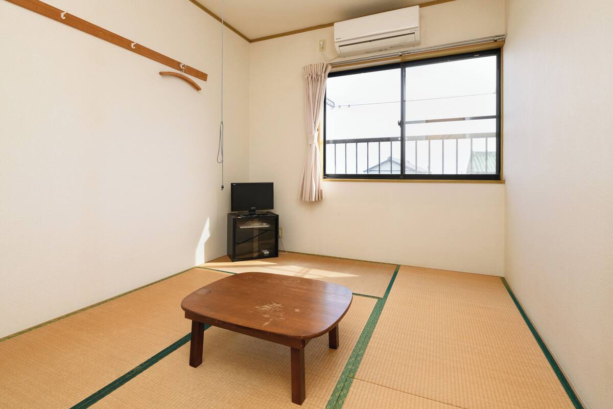 Ein Zimmer mit Tatami-Boden, einem Holztisch, einem Fernseher und einem Fenster mit Blick auf Gebäude.