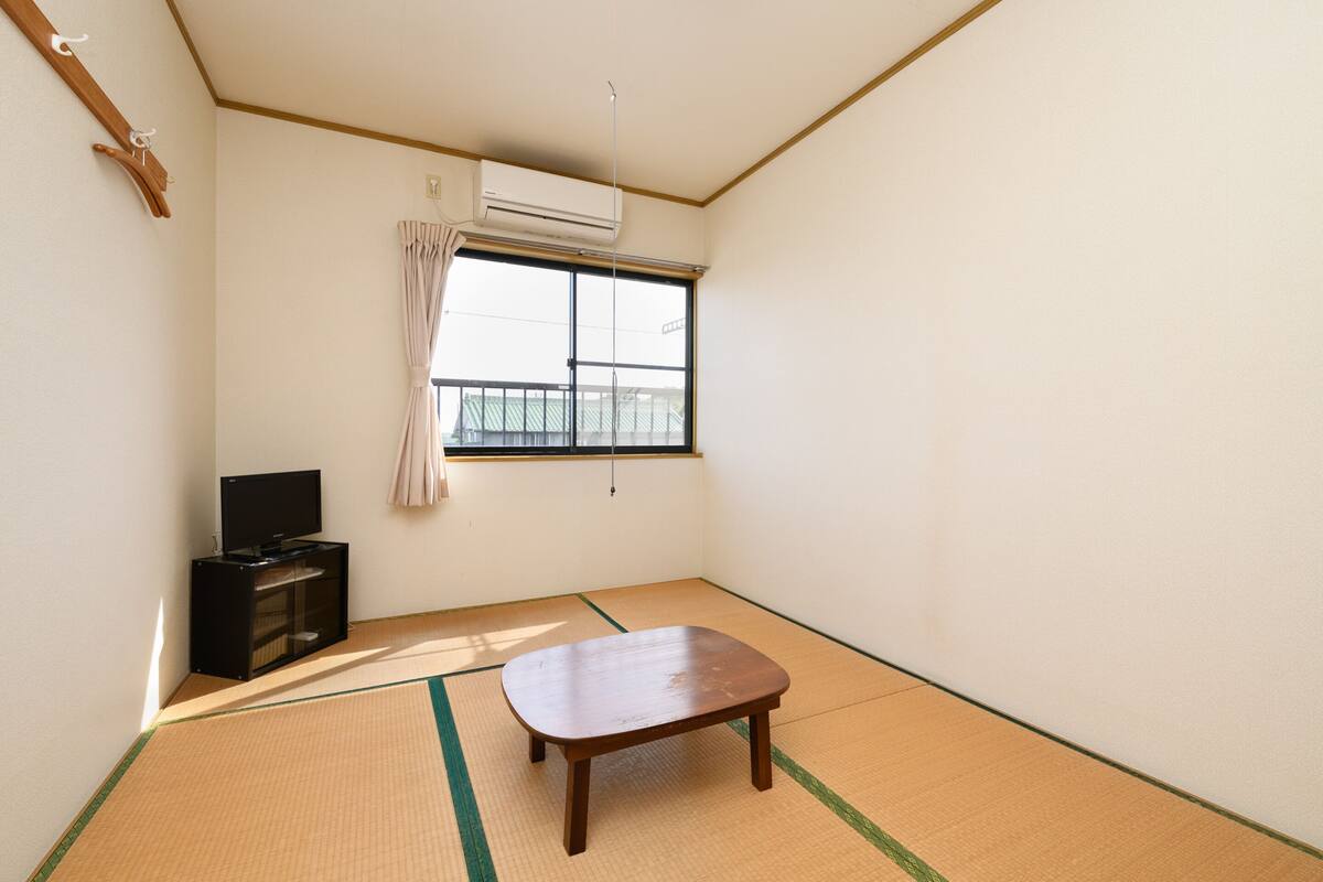 Ein Zimmer mit Tatami-Böden, einem kleinen Holztisch, einer schwarzen Unterhaltungseinheit und einem Fenster mit Blick auf ein Gebäude.