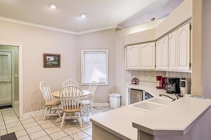 Una cocina compacta con muebles blancos, una mesa de comedor con sillas, una ventana con persianas y un cubo de basura.