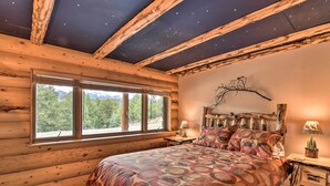 Ein Schlafzimmer in einer Blockhütte mit einem großen Bett, Holzbalken, einem Fenster mit Blick auf Bäume und einer rustikalen Wanddekoration aus Ästen.
