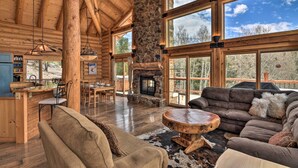 A spacious log cabin interior with a stone fireplace, wooden furniture, and large windows offering views of the outdoors. - Spacious, Luxe Cabin w/ Mtn Views, Sauna & More! (Villa Grove)