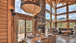 A spacious log cabin with a dining area, a stone fireplace, and a large window overlooking a mountainous landscape. - Spacious, Luxe Cabin w/ Mtn Views, Sauna & More! (Villa Grove)