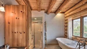 Ein rustikales Badezimmer einer Holzhütte mit Holztür, Dusche, Badewanne und einem Fenster mit Blick ins Freie.