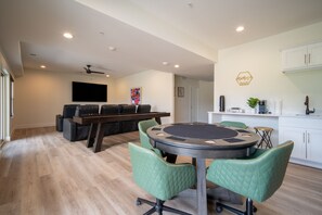 A modern living room with a pool table, green chairs, a flat-screen TV, and a kitchen area with cabinets and a coffee machine.