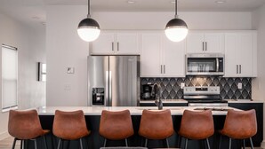 A modern kitchen with white cabinets, stainless steel appliances, and a black and white backsplash. There are six brown bar stools around the island. - Gorgeous Reunion Lodge with 2 Decks + Patio, Resrot Amenities, Ticket & Golf Deals! (Branson)