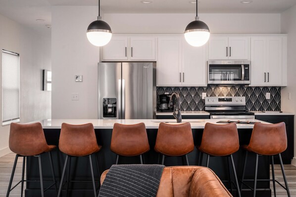 A modern kitchen with white cabinets, stainless steel appliances, and a black and white backsplash. There are six brown bar stools around the island.
