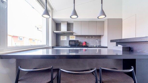 Una cocina moderna con una isla grande, tres lámparas colgantes, un microondas, un hervidor rojo y vistas al exterior.