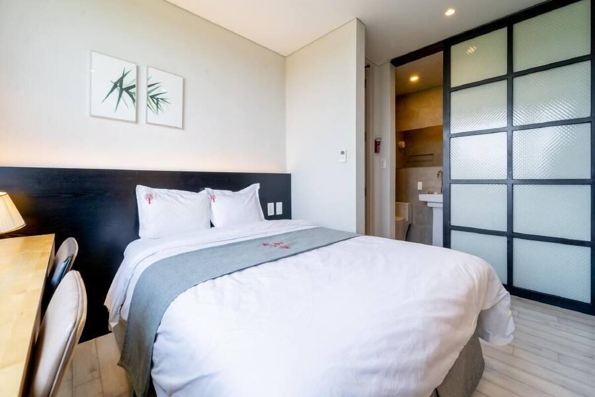 Habitació d'hotel amb un llit, un escriptori i vista a un bany a través d'una porta de vidre.