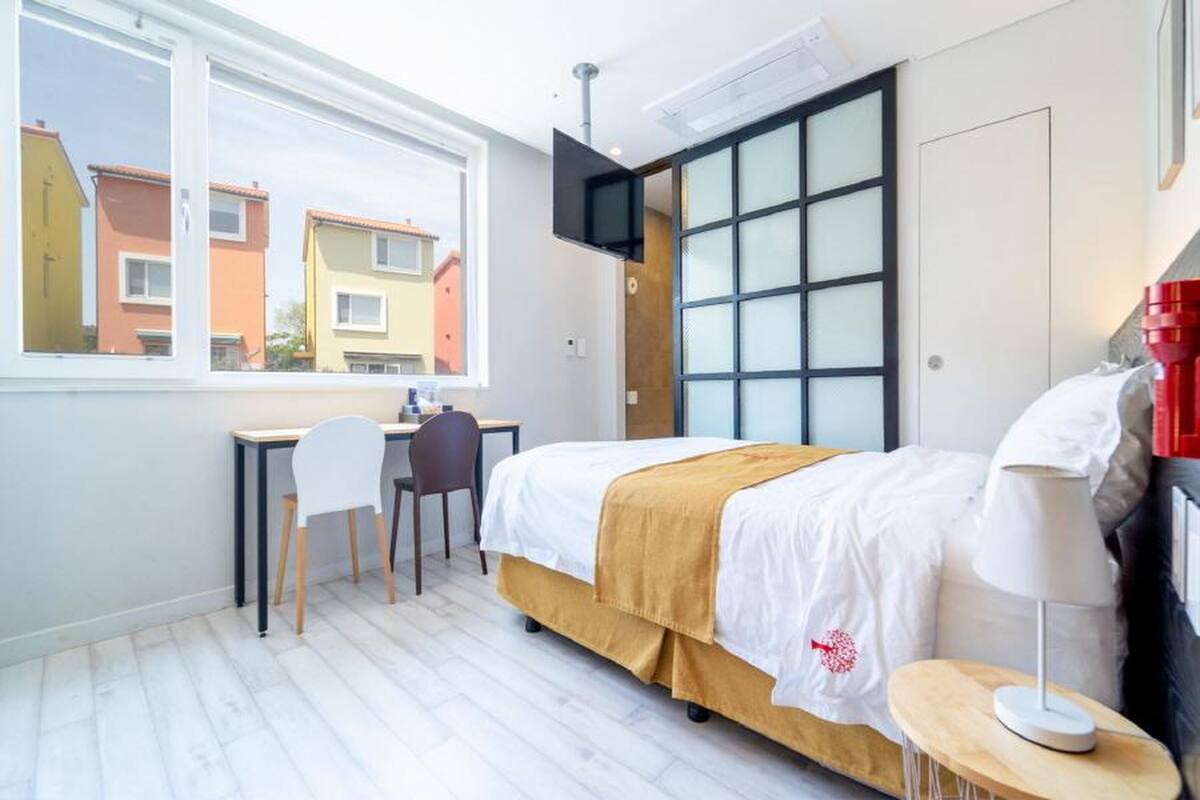 Un dormitori amb un llit, una taula de treball, una cadira, un televisor fixat a la paret i una finestra que ofereix vista a edificis veïnals.