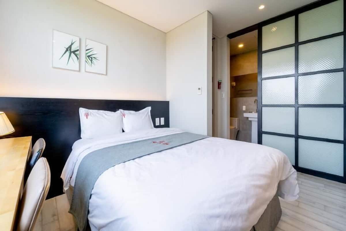 A hotel room with a bed, a desk, and a view of a bathroom through a glass door.