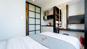 Habitación de hotel con cama, televisor montado en la pared, una pequeña cocina y puerta corrediza.