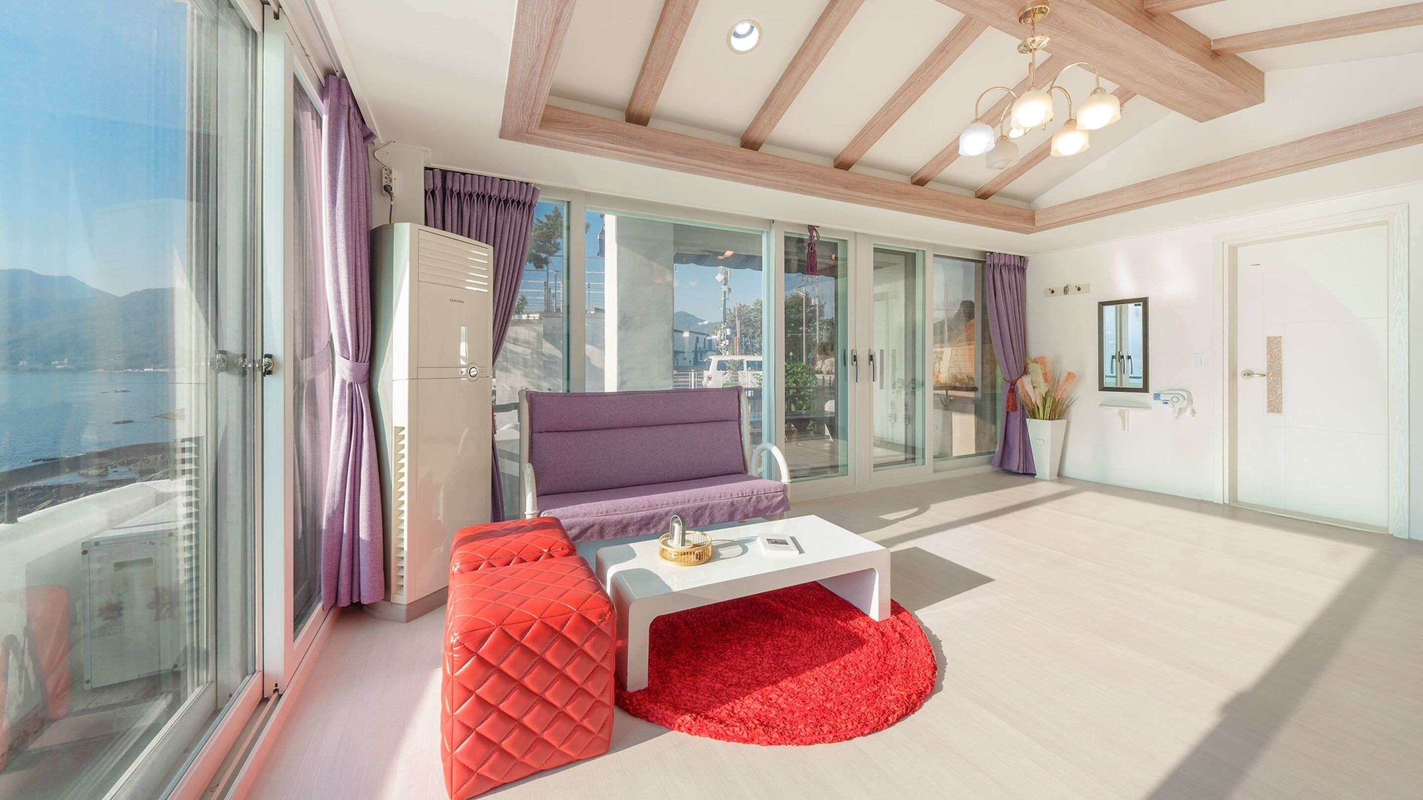 A modern living room with a sofa, a red ottoman, a white coffee table, and a view of the sea through large windows.
