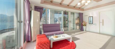 A modern living room with a sofa, a red ottoman, a white coffee table, and a view of the sea through large windows.